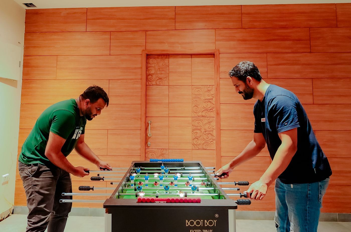 Two people play foosball in a room with brick walls and a high ceiling - Nirantara Resort
