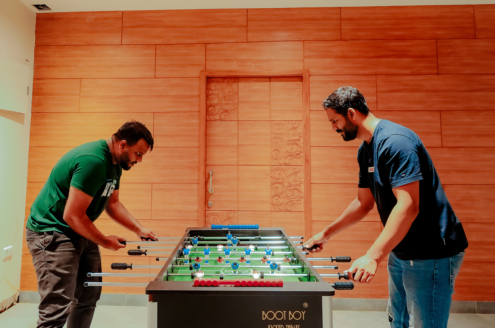 Two people play foosball in a room with brick walls and a high ceiling - Nirantara Resort & Spa.
