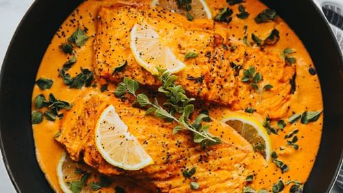  a fish dish featuring orange coloured gravy garnished with some greens.