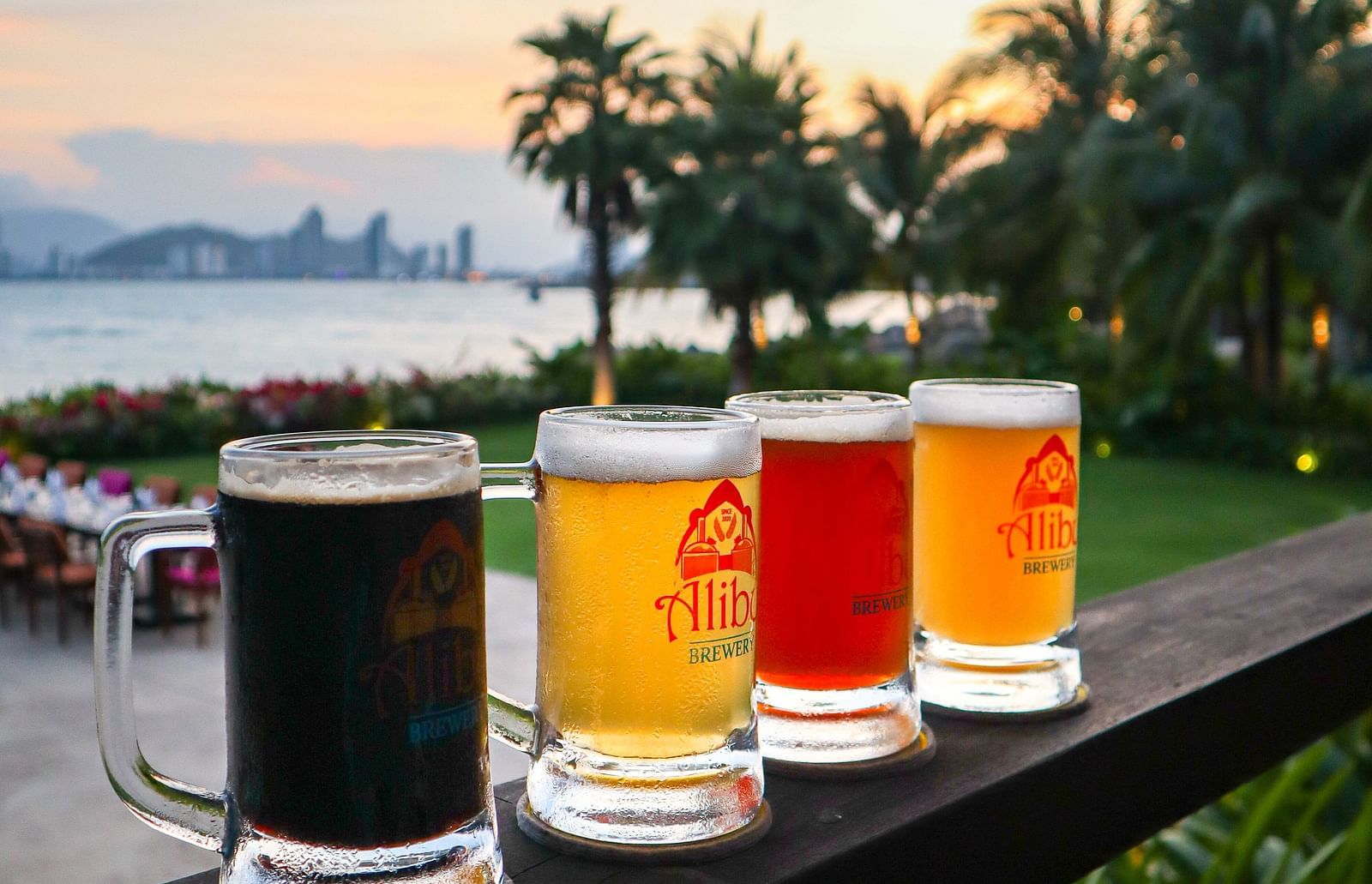 An array of drinks placed on a wooden railing near a beach - Alibu Resort Nha Trang