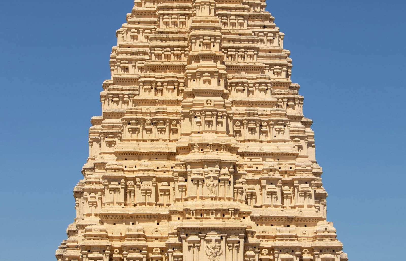 A tall, ancient Hindu temple tower (shikhara) with intricate carvings, shown from a low angle against a clear blue sky.