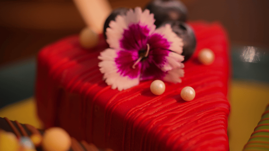 A slice of bright red cake is adorned with a purple and white flower, blueberries, and pearl-like sprinkles on top. The mood is vibrant and elegant.