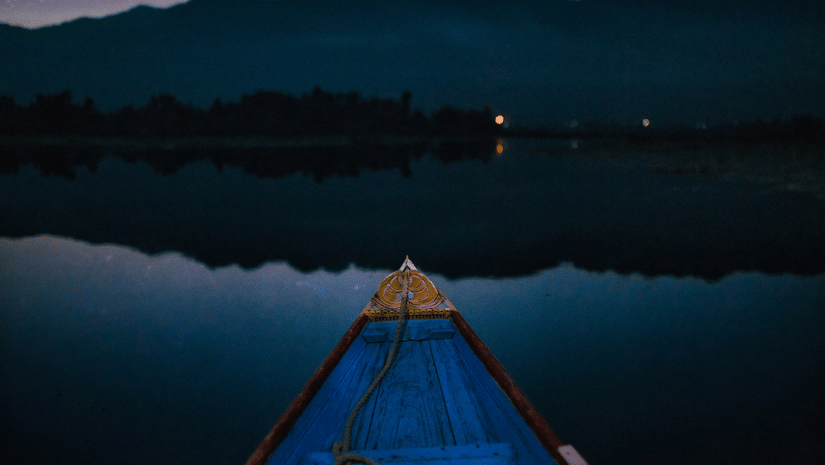 Wooden boat on still lake at night, pointing toward star-filled sky and mountain silhouettes reflected on calm water surface below