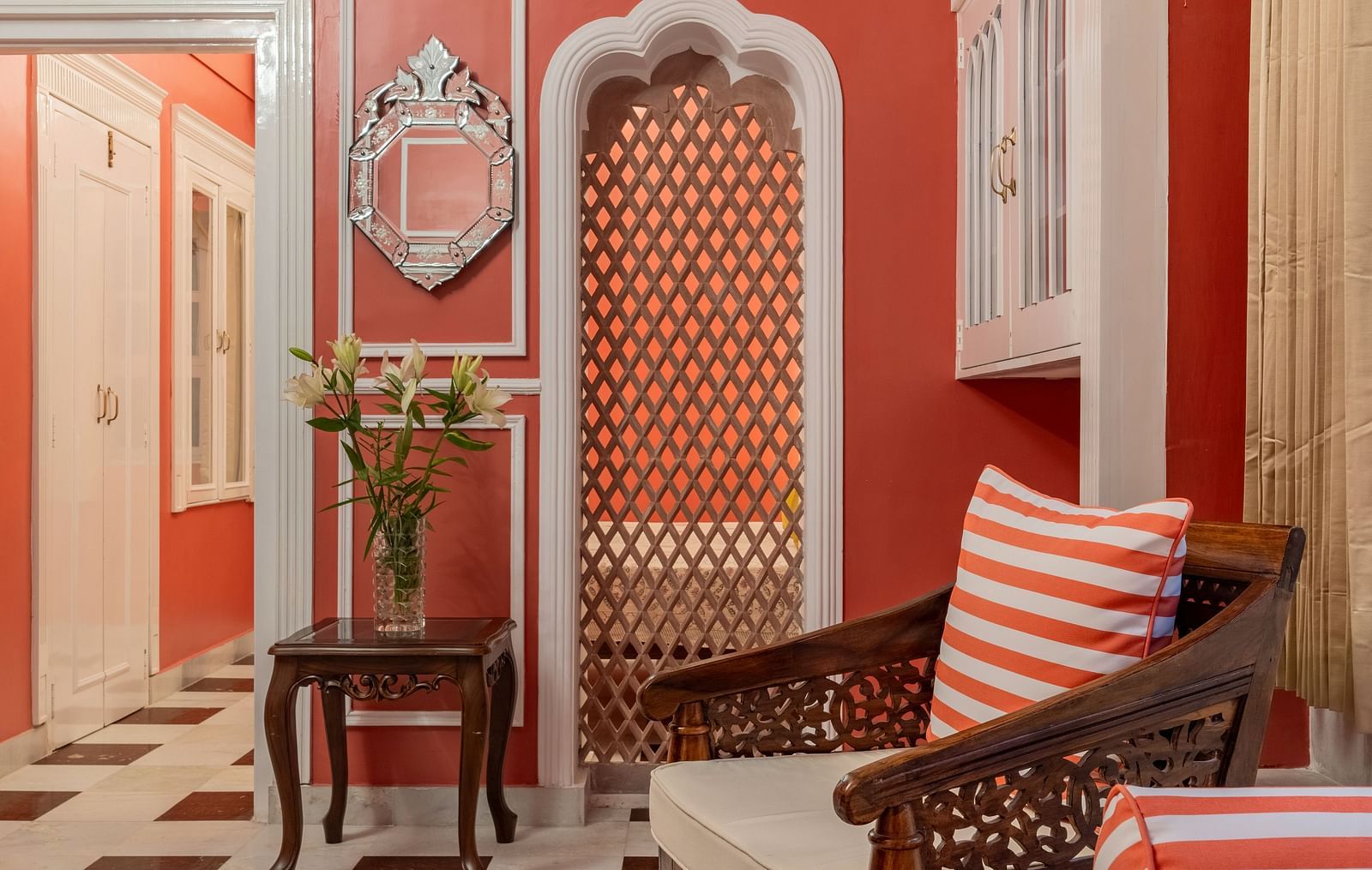 A hallway with red walls, white trim, a checkered floor, a wooden chair with a striped cushion, and an arched doorway | Dera Rawatsar, Jaipur