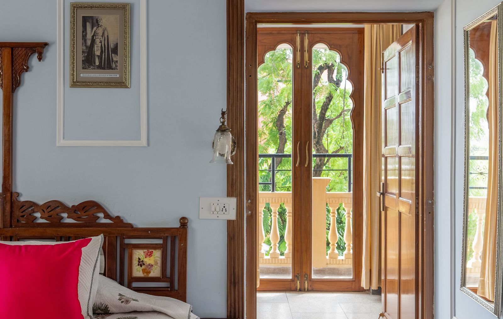 A serene bedroom at Dera Rawatsar, Jaipur, features a comfortable bed, a patterned rug, and an ornate wooden doorway leading to a bright space.
