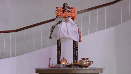 A small idol draped in white cloth with an orange garland, placed on a wooden table with ritual items, near a staircase railing on the Benzz Park property.