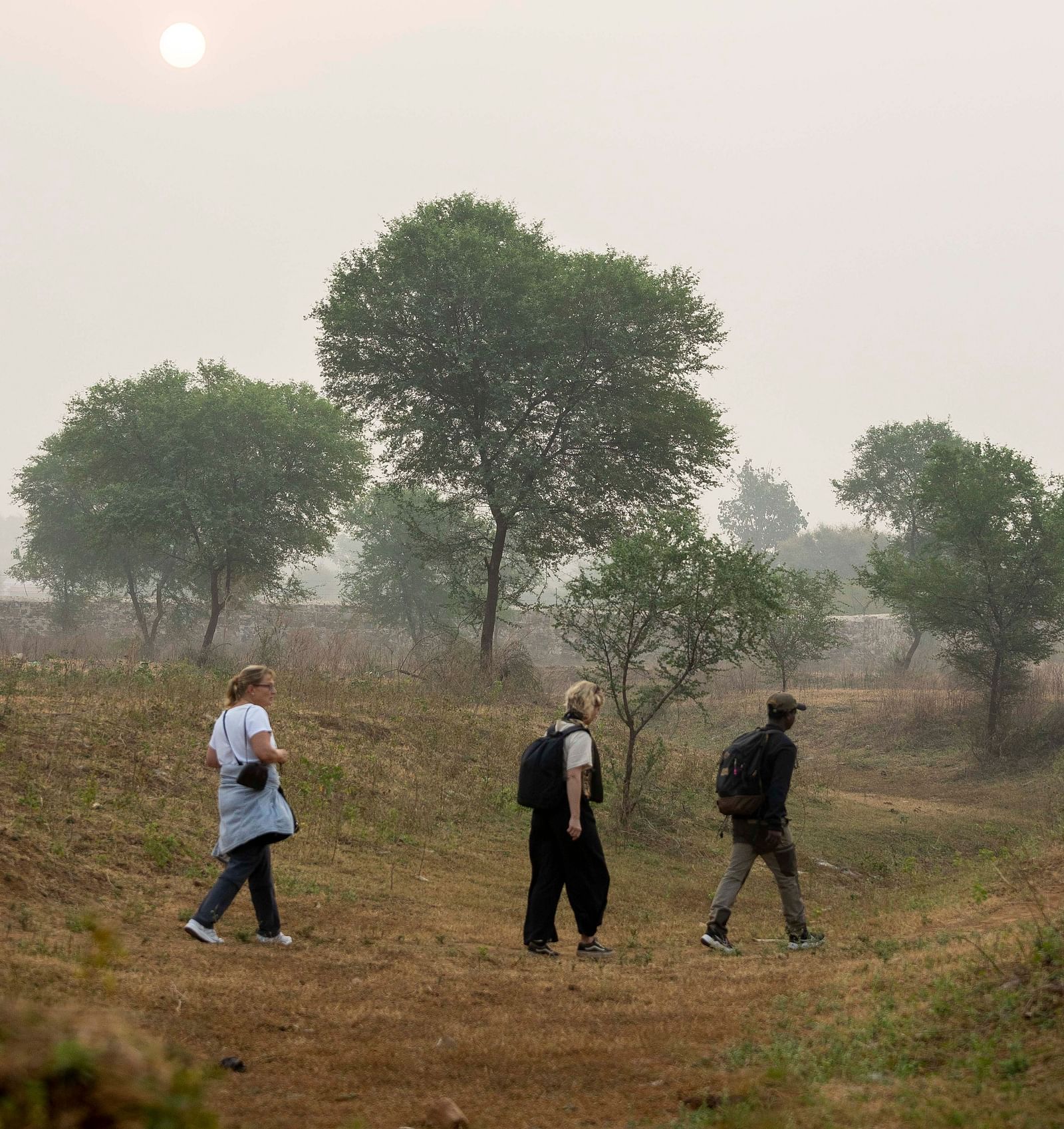 An image of three people walking through an arid land with few trees in the background - An image of two women with a tourist guide - Utsav Camp Sariska