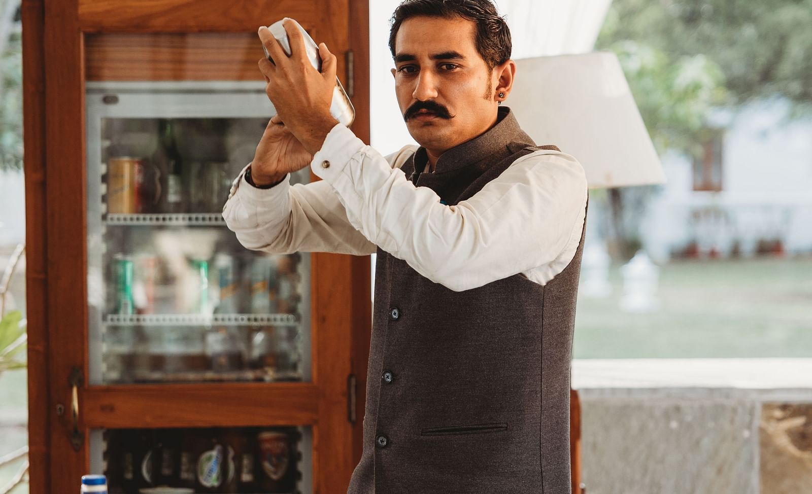 A person standing behind a bar counter with several bottles of alcohol lined up in a row- Shahpura Bagh.