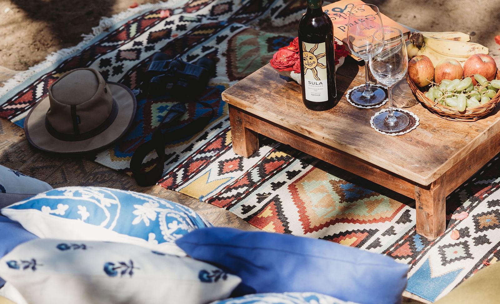 A picnic setup on a sandy surface with a small wooden table, pillows, a blanket, a basket with flowers, and a hat- Shahpura Bagh. 