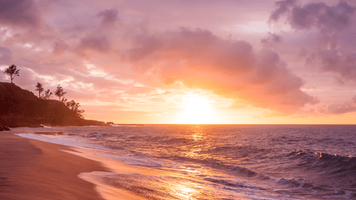 A beach seashore with layers of waves seen during a vibrant sunset under a partially cloudy yet dramatic sky.