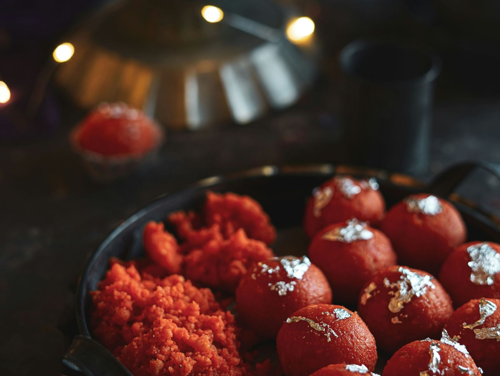 A close-up of a platter containing bright orange Ladoo sweets, some decorated with silver leaf, with bokeh lights in the background.