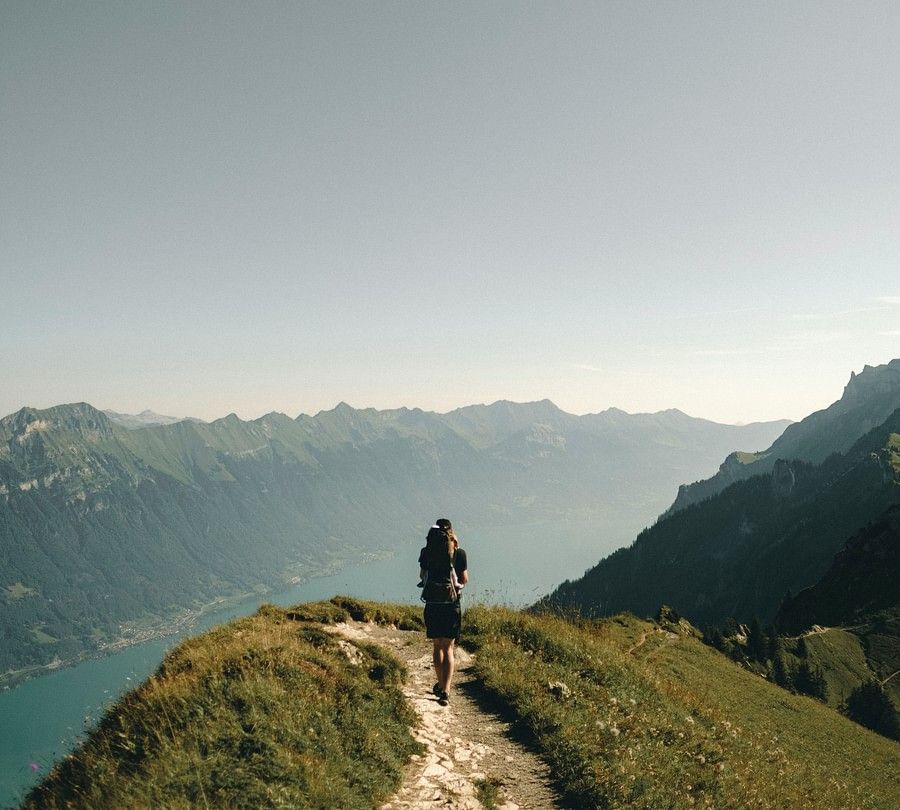 A lone hiker follows a narrow trail on a mountain ridge with dramatic views of rugged terrain and distant peaks under a bright sky.
