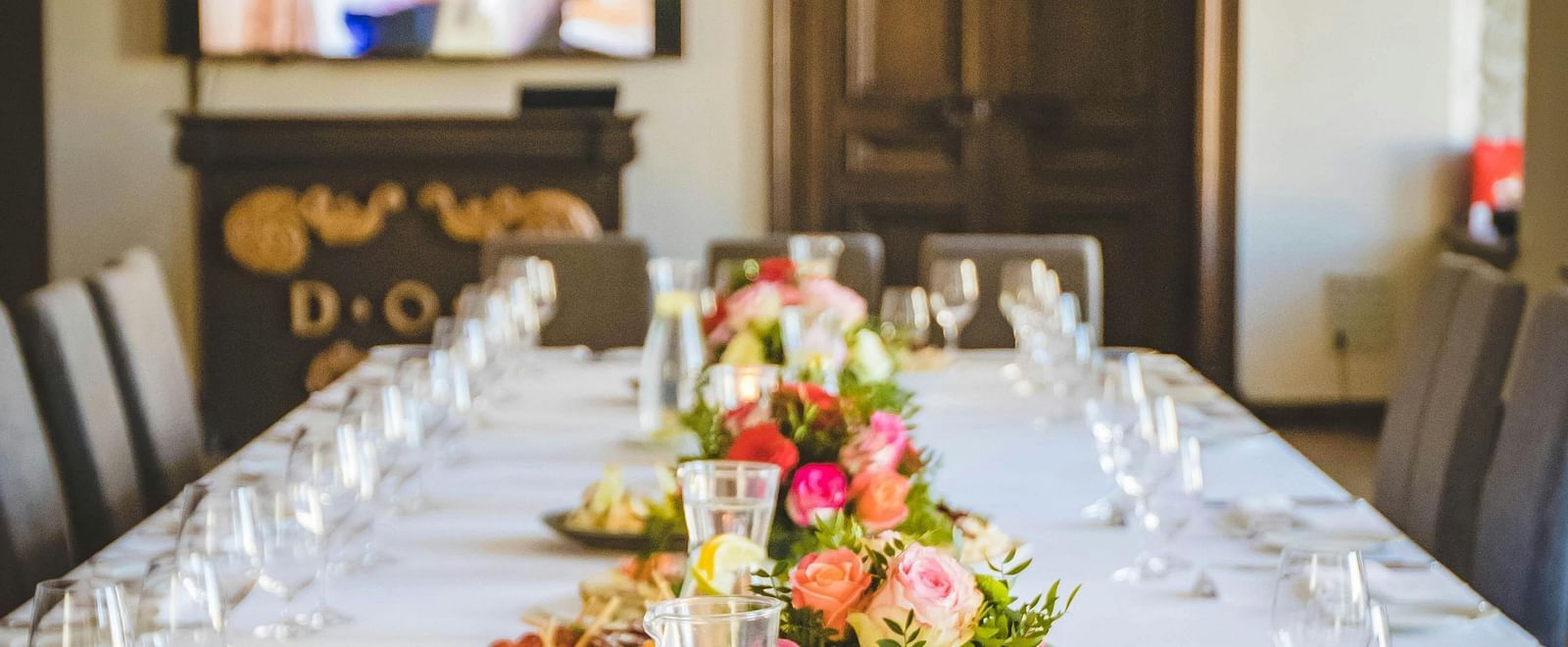 A long dining table decorated with flowers in the middle along with shiny glassware and cutlery set on the table.
