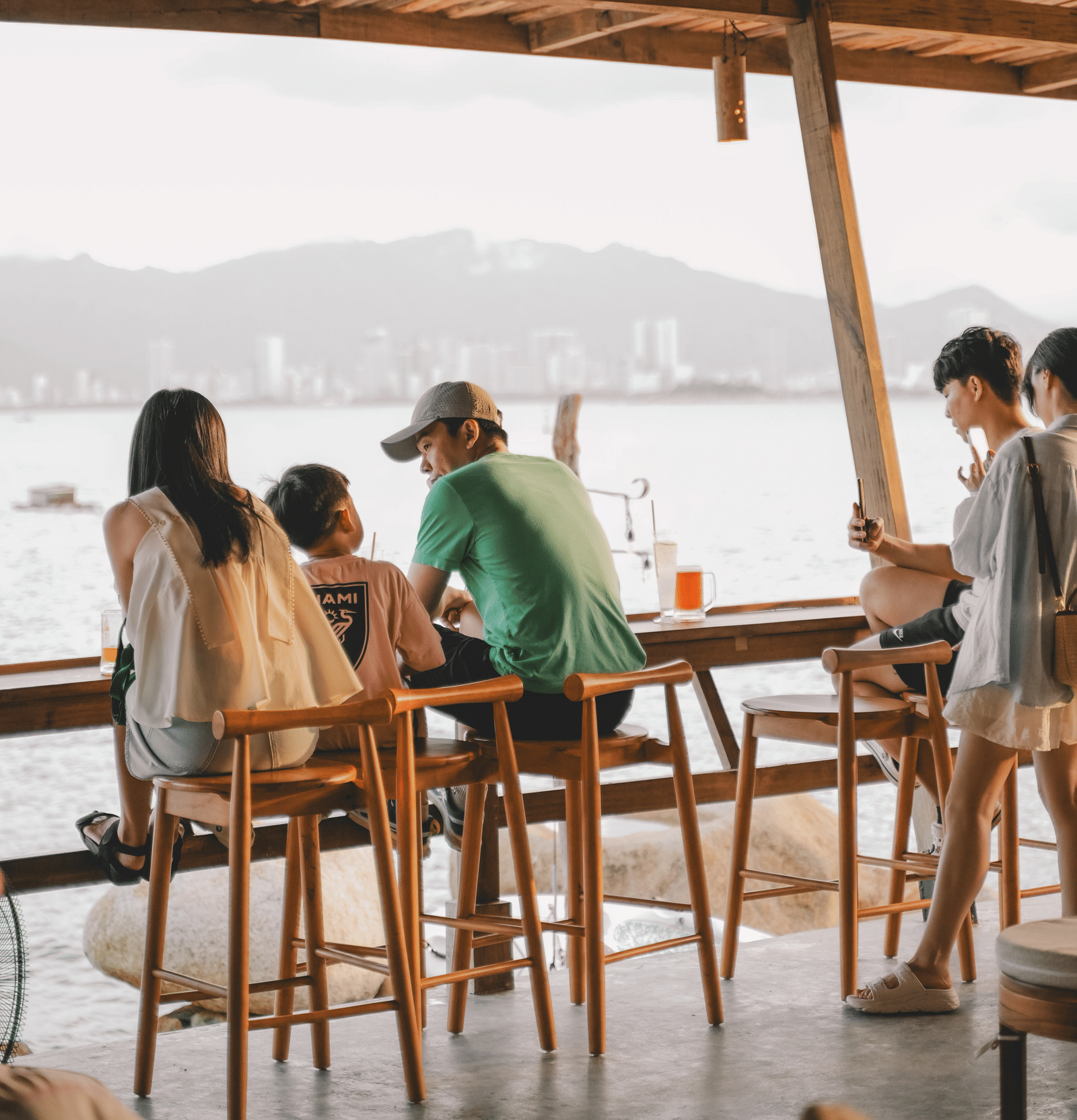 People sitting a wooden furnished restaurant which has ceiling lights and a river view.
