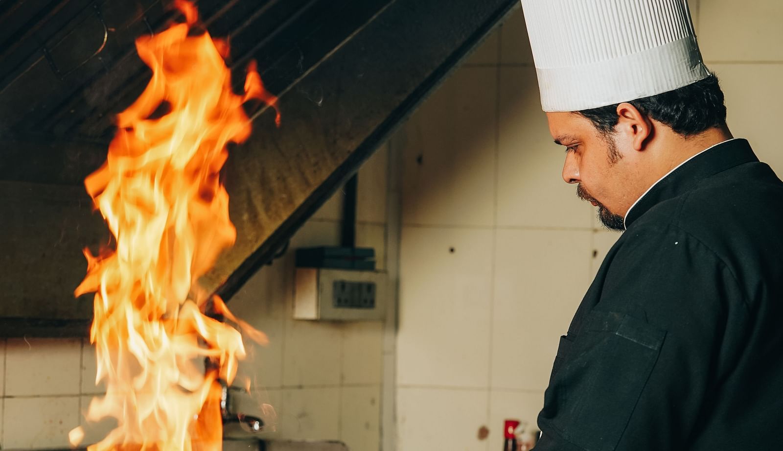 A chef at Orsino Hotel and Spa in tall white hat and black uniform is cooking in a wok with large flames rising.