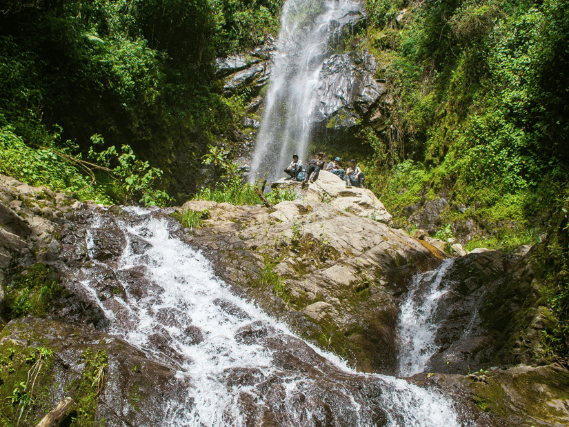 An image of a waterfall cascading through the rocks amidst the lush greenery of the forest with people enjoying the clean stream of water
