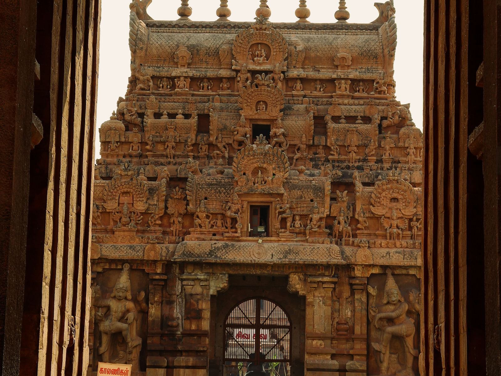 A view through an ornate stone archway reveals a towering, intricately carved temple gateway with people walking around.