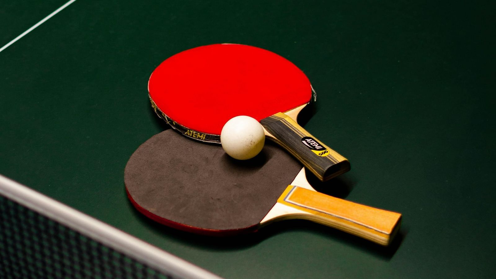 Two table tennis paddles, one red and one black, resting on a green ping pong table with a white ball placed on top.