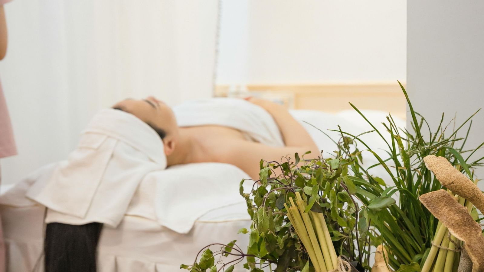 Woman relaxing on a spa bed with white towels, beside a basket of fresh herbs and a lit candle creating a calming ambiance.