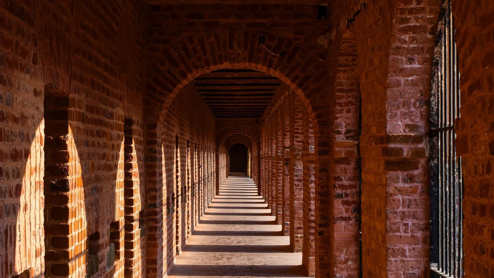 A corridor featuring walls made of red bricks with sun rays falling on the wall shaped by the arched pillars.