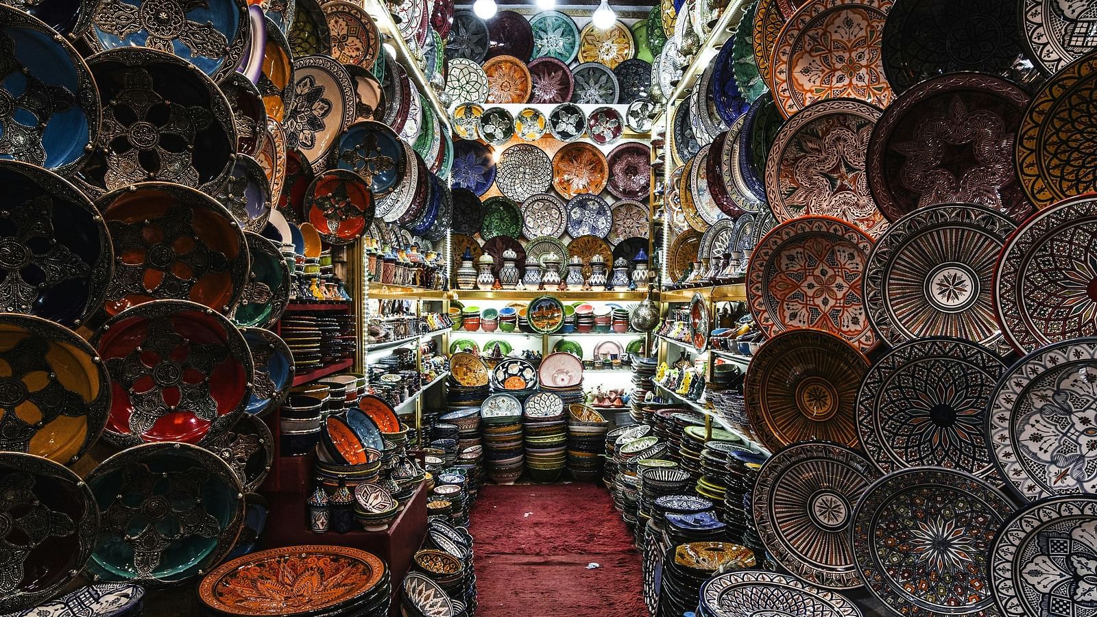 A narrow aisle in an Oriental shop, filled with colorful bowls, plates, and hanging lamps, creates a vibrant and immersive shopping experience.