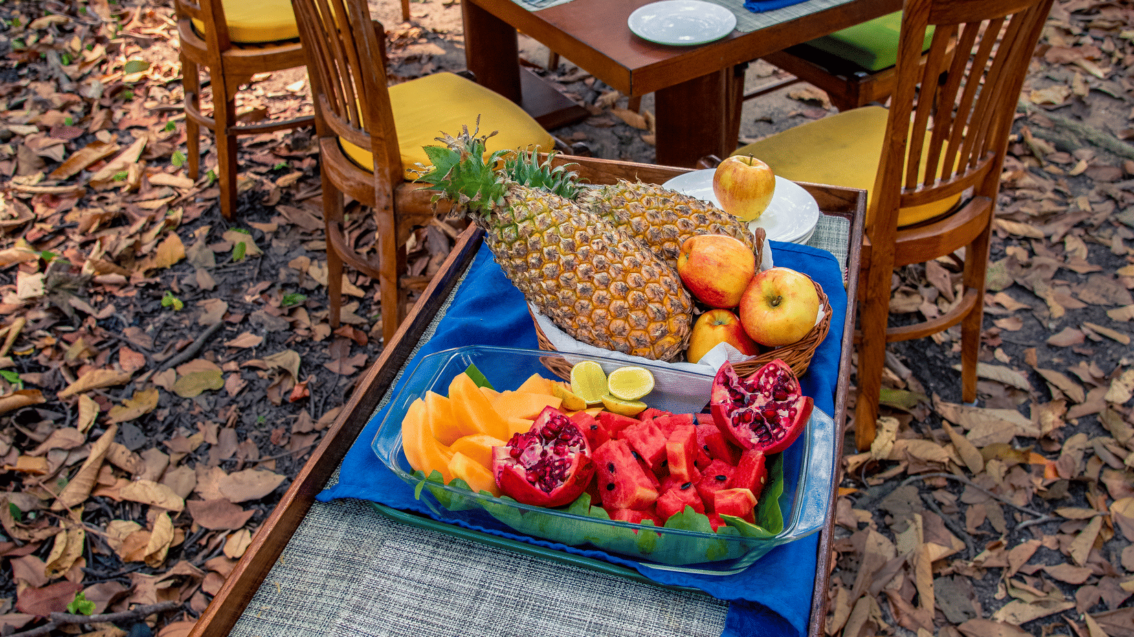 A wooden dining table with chairs and a separate tray set up outdoors in a forested area covered in fallen leaves.
