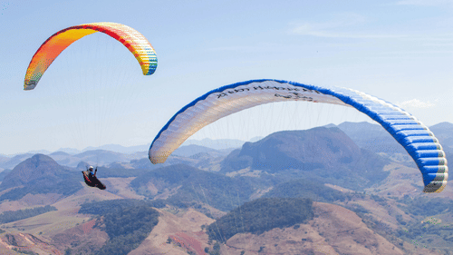 2 people fly Vagamon paragliding together above rolling mountain terrain, their colourful canopies contrasting with the rugged landscape and expansive blue sky