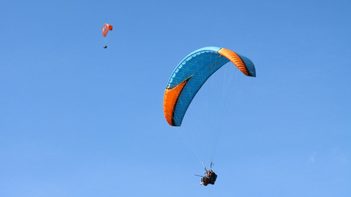 A colourful paraglider drifting high against a clear blue sky, with tiny landscape details visible far below the open canopy