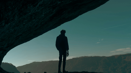 A lone figure stands silhouetted beneath a vast rock overhang.
