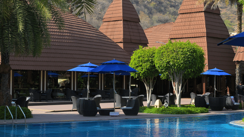 Serene outdoor swimming pool surrounded by lounge chairs and blue umbrellas, with traditional architecture and palm trees in the background, set against a backdrop of hills - Ananta Spa and Resort, Pushkar.