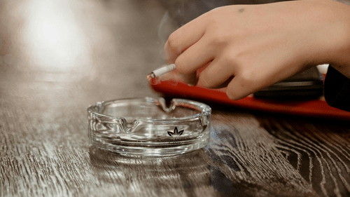 a person holding a cigarette over a crystal ash tray kept on a wooden counter top 