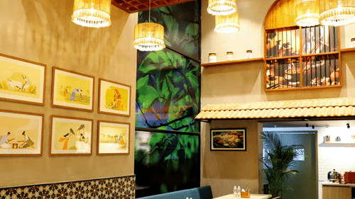 The interior of a casual restaurant or cafe with wooden tables, booth seating, hanging light fixtures, and framed pictures on the walls.