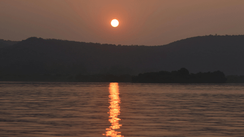 The sun sets above distant hills, casting a reflection across the surface of a body of water.