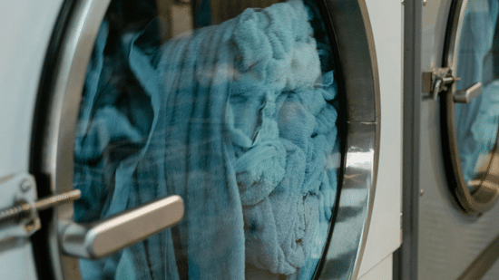 A close-up side view of a commercial washing machine with light blue fabrics visible through its circular glass door.
