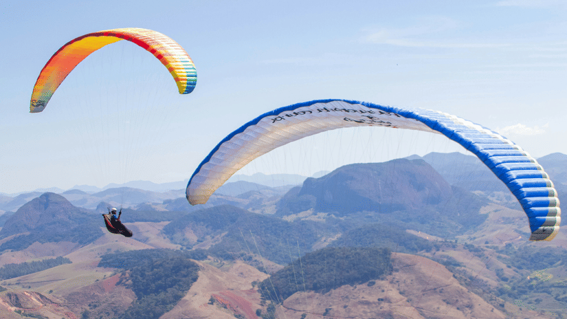 2 people fly Vagamon paragliding together above rolling mountain terrain, their colourful canopies contrasting with the rugged landscape and expansive blue sky
