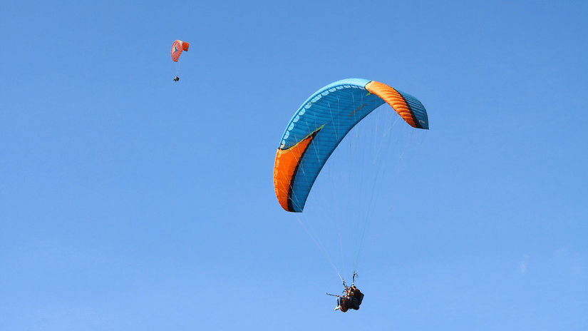 A colourful paraglider drifting high against a clear blue sky, with tiny landscape details visible far below the open canopy