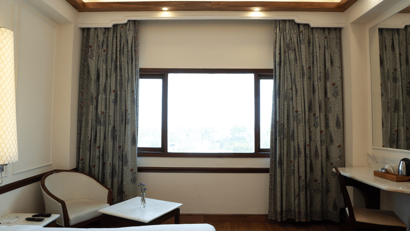 Hardwood floors lead toward a window and a desk area illuminated by recessed ceiling lights in the Deluxe King room at Clarks Amer, Jaipur.