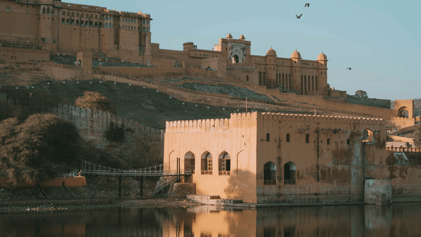 A panoramic view of Amer Fort rising on a hill, reflected in the waters of Maota Lake, and under clear blue sky.