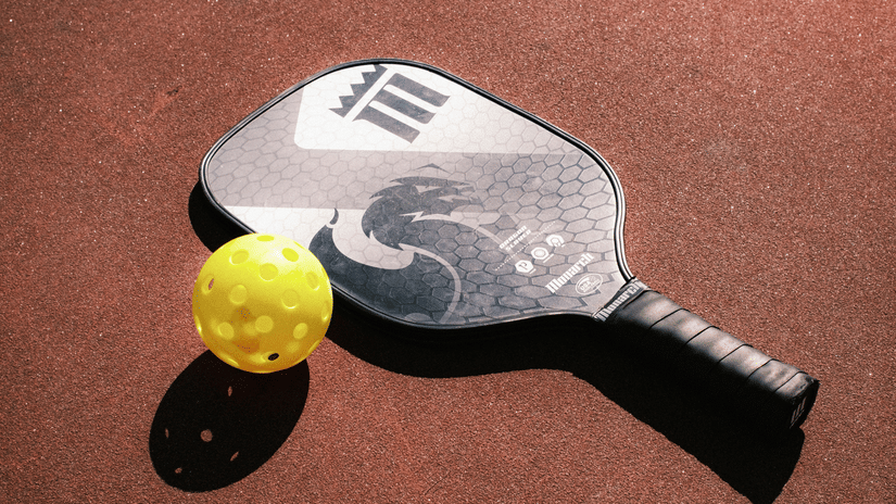 Black pickleball paddle and yellow ball lying on a textured court floor in soft spotlighting