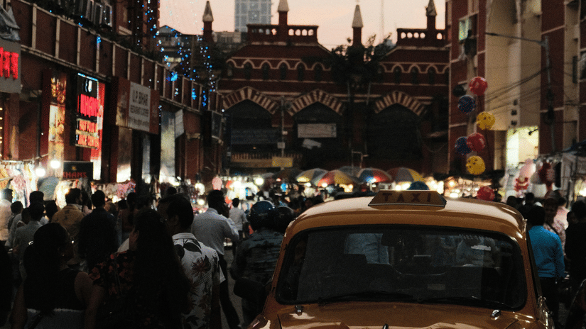 A street market in Kolkata with numerous people walking on the road and a yellow taxi