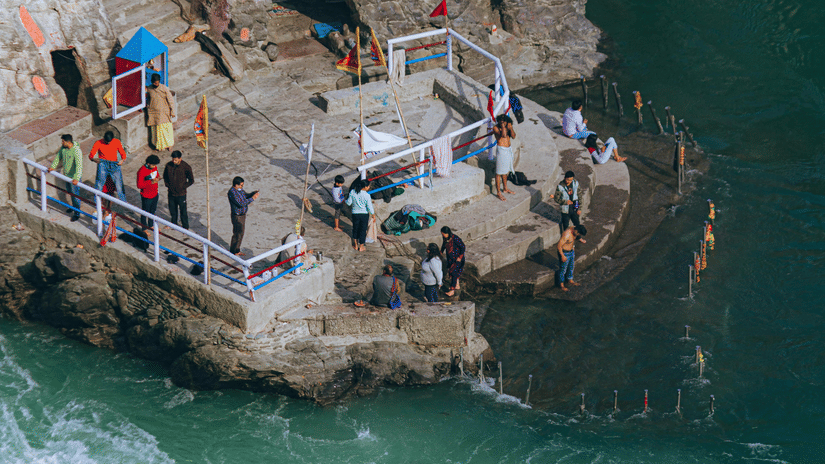 A high-angle vertical shot of a stone ghat with people gathered near the swirling green waters of a fast-flowing river.