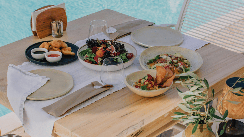 A wooden table is set with plates of food next to a swimming pool.