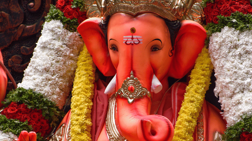 A vibrant Ganesha idol adorned with a crown and garlands stands against an ornate backdrop during a colourful festival celebration