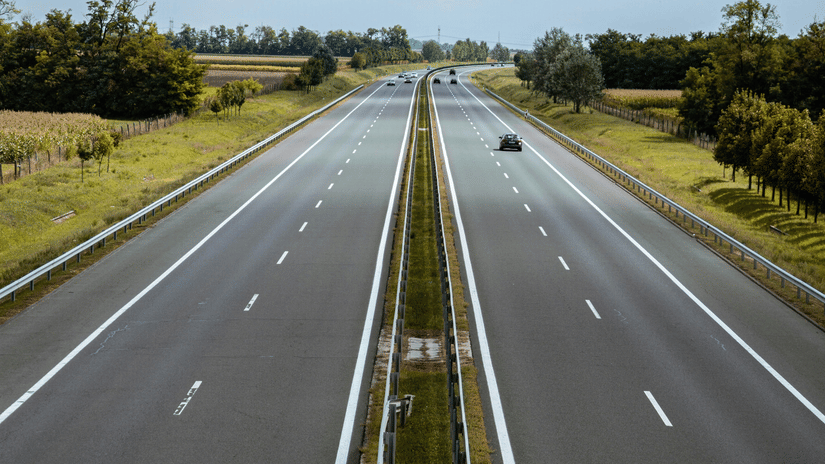 A view atop of a four lane highway divided by a small lawn, cars travelling on the road and scattered trees on either side of the road.