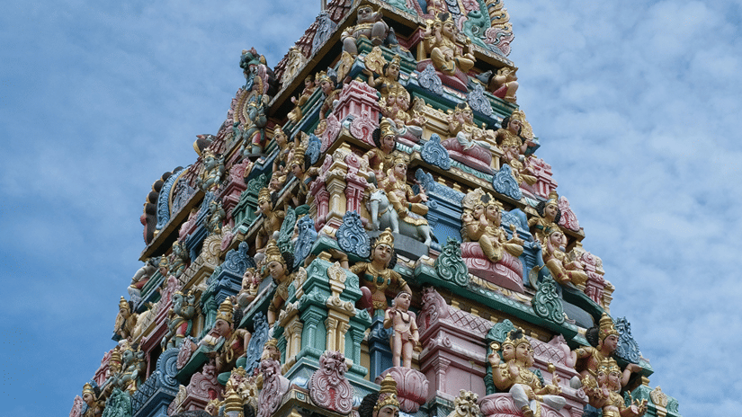 A view from below of Pillayarpatti Ganapathi temple's facade with numerous intricate small deities carved into the gopuram and blue sky in the background..