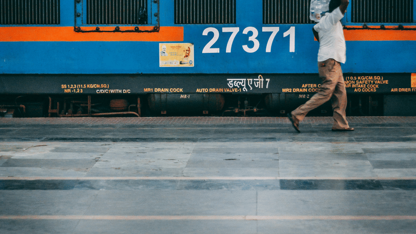 A person walking next to a train on a train station on a platform carrying packaged drinking water with a building in the background.