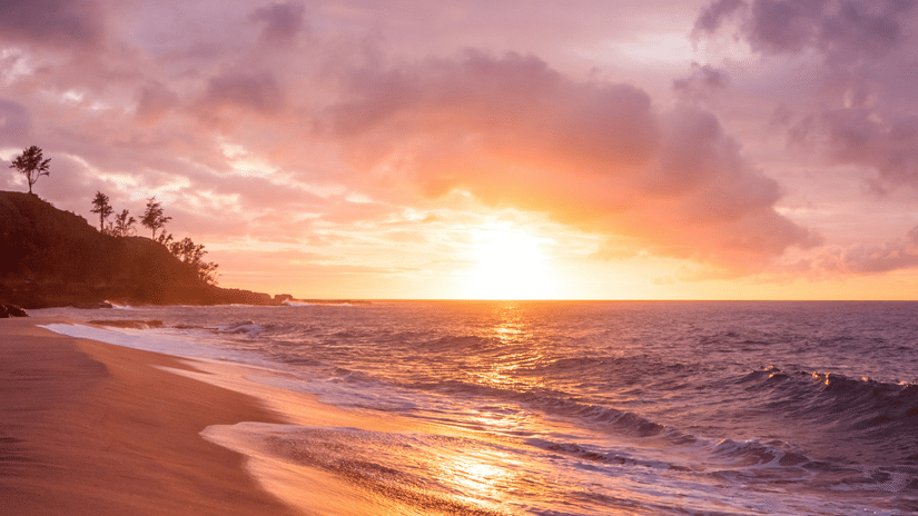 The shore of a beach during sunset, with waves reflecting the warm colours of the sky.