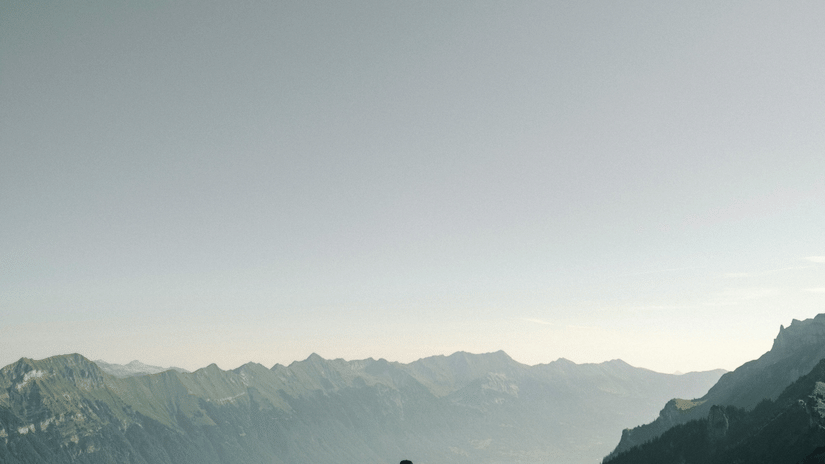 A solitary hiker with a backpack walks away from the camera on a grassy, dirt path along a ridge overlooking a vast valley and mountain range under a pale sky.