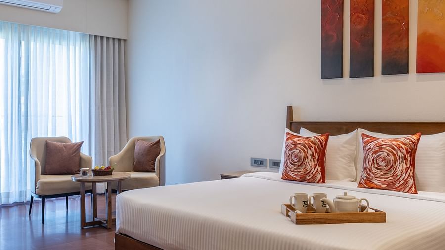 An image of a bedroom with a king size bed where tea tray is placed, a flat TV screen on top of a table counter top, a seating area at one corner, and a  large window at one side