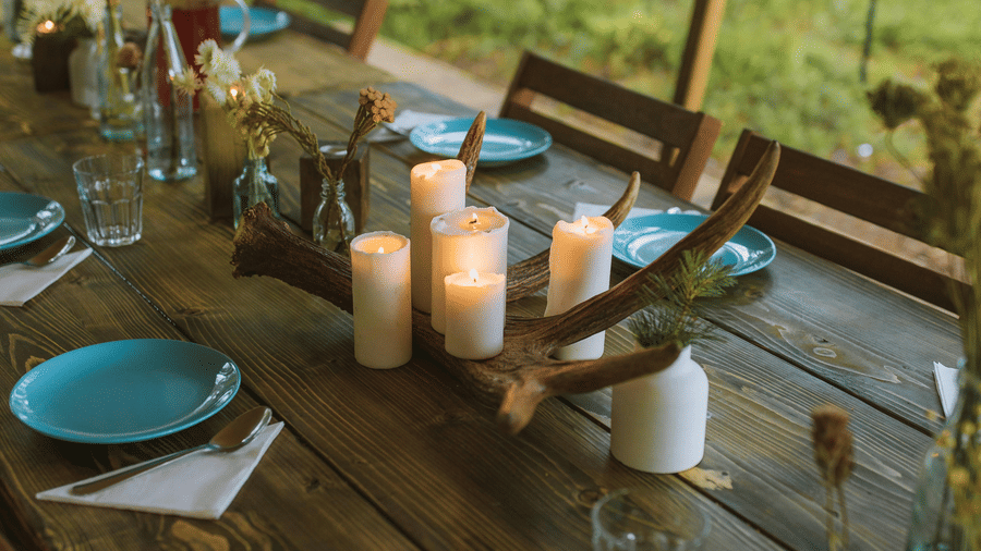 A rustic wooden table set for an outdoor meal under a tent, featuring plates, candles, a dried antler centrepiece, and a forest background.
